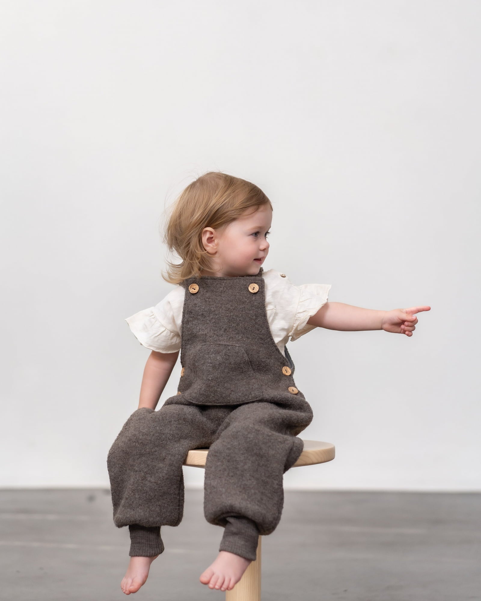 Kind in Wollwalk Latzhose in Farbe Nuss 
auf Hocker