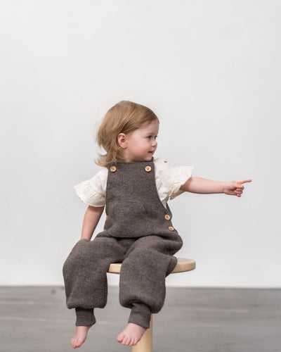 Kind in Wollwalk Latzhose in Farbe Nuss 
auf Hocker